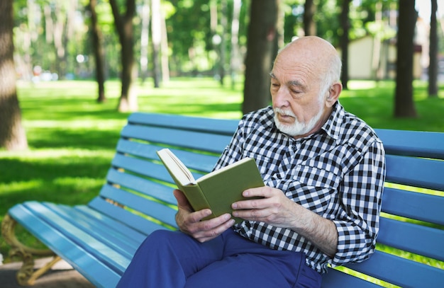 Libro di lettura dell'uomo anziano all'aperto. Uomo anziano casual che si riposa sulla panchina nel soleggiato parco estivo, copia spazio