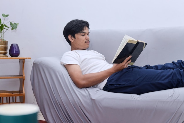 Libro di lettura dell'uomo asiatico a casa rilassante su un divano