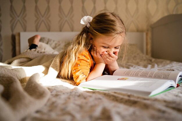 Libro di lettura della bambina sveglia che si trova sul letto nello stile di vita interno domestico