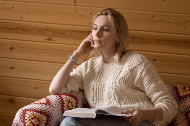 Libro di lettura della donna bionda e pensiero seduto sul divano di casa al fondo della parete di legno