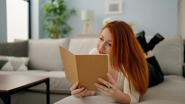 Libro di lettura della giovane donna della testarossa che si trova sul sofà a casa