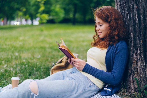 Libro di lettura della giovane signora di bell'aspetto e sorridente seduto sul prato nel parco