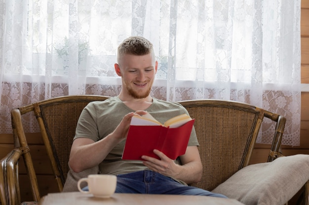 Libro di lettura giovane uomo sorridente con copertina rossa sulla panca di vimini in casa di campagna in legno