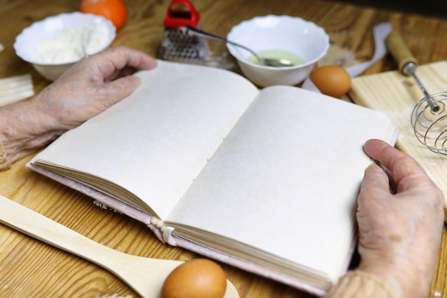 Libro di ricette aperto nelle mani di una donna anziana davanti a un tavolo con utensili