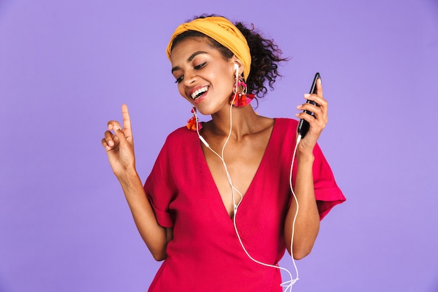 Lieta donna africana in abito e auricolari ascoltando musica con gli occhi chiusi sul muro viola