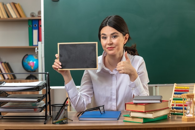 Lieto di mostrare pollice in su giovane insegnante femminile seduta a tavola con strumenti scolastici che tengono mini lavagna in classe