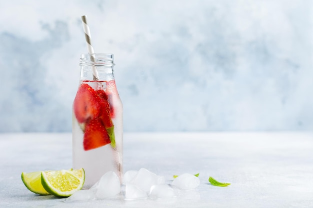 Limonata estiva con ghiaccio, fragole, menta e lime in una bottiglia e un tubo di carta su uno sfondo grigio cemento. Concetto di bere sano