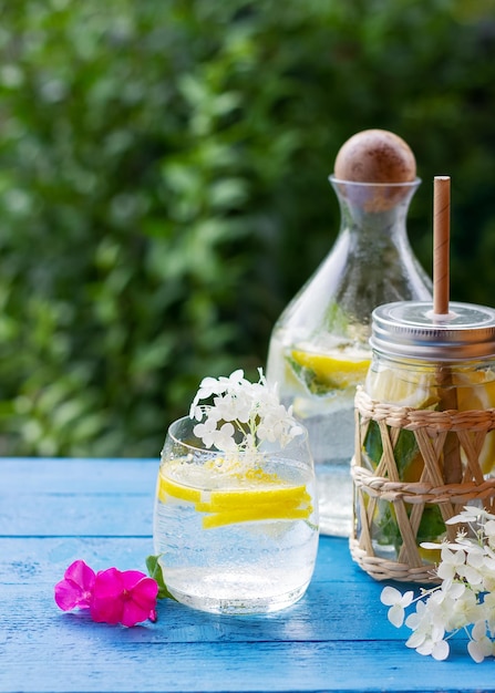 Limonata fatta in casa con limone e cubetti di ghiaccio in una brocca di vetro sul tavolo di legno blu con fiori bianchi e rosa