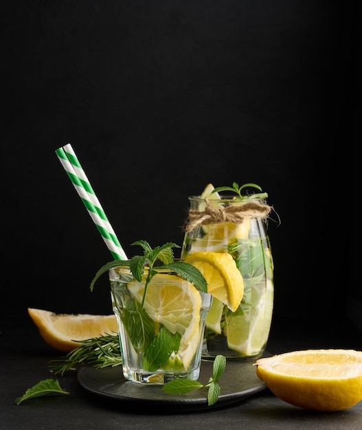 Limonata in un bicchiere trasparente con rametti di rosmarino limone lime e foglie di menta su sfondo nero