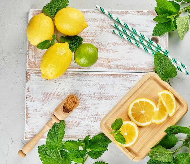 Limoni interi gialli maturi e lime, zucchero di canna e un mazzetto di menta fresca su una tavola di legno bianca