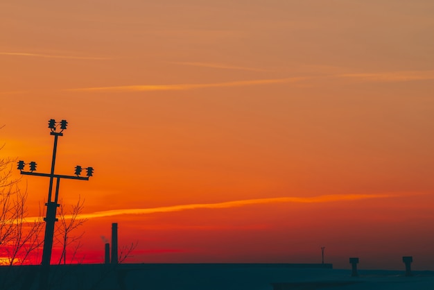 Linee elettriche sopra il tetto all'alba. Siluette del mucchio con i fili fra smog su alba. Cavi ad alta tensione sul caldo cielo giallo arancio. Industria energetica al tramonto. Alimentazione elettrica della città. Nebbia sfondo urbano.