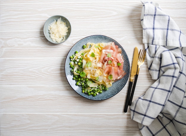 Linguine con besciamella, piselli, parmigiano, prosciutto e menta fresca in un piatto blu su legno bianco