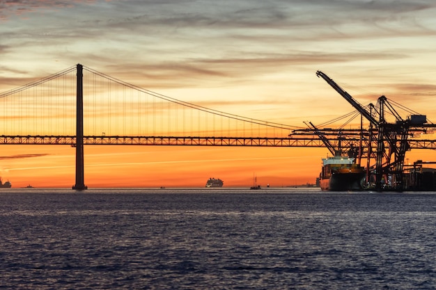 Lisbona e il ponte di aprile al tramonto gru industriali che lavorano sull'acqua