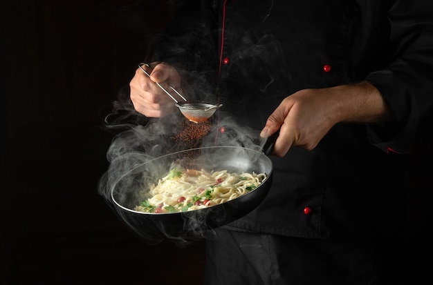 Lo chef aggiunge spezie agli spaghetti in una padella calda al vapore Ricetta o menu per ristorante o hotel su sfondo nero