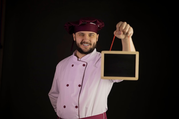 Lo chef barbuto bello positivo in uniforme tiene il bordo di gesso in bianco