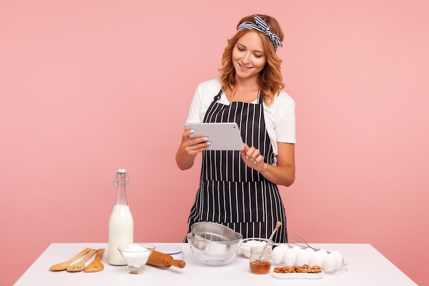 Lo chef casalinga cuoco pasticcere o fornaio alla ricerca di una nuova ricetta in internet in piedi con tablet in mano vicino al tavolo con prodotti per la cottura Studio indoor girato isolato su sfondo rosa