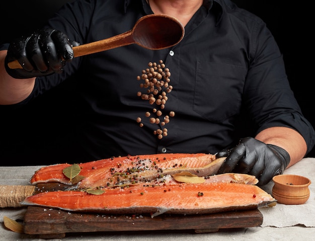 Lo chef in abiti neri e guanti in lattice nero versa pimento su una carcassa affettata di pesce salmone fresco