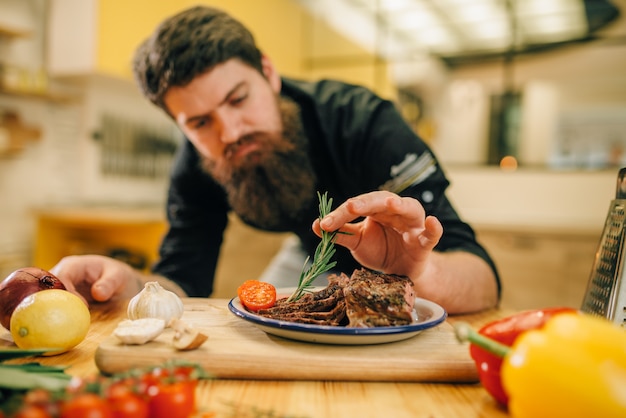 Lo chef maschio decora con erbe aromatiche fette di carne in un piatto, cucina sullo sfondo. Uomo che prepara carne di manzo con verdure sul piano di lavoro