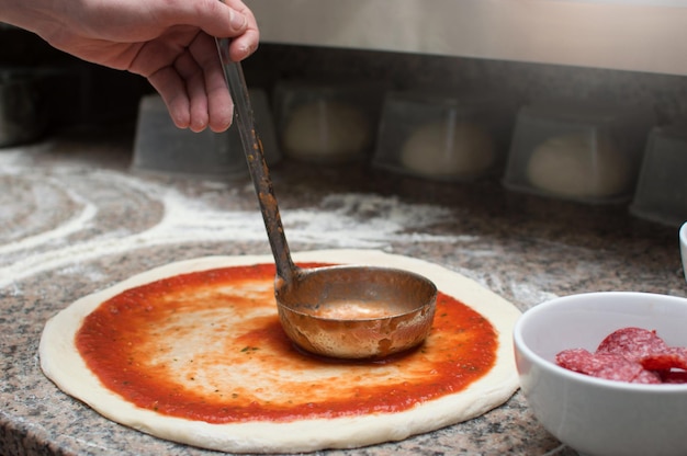 Lo chef maschio spalma la pizza piccante con salsa rossa Preforma di pasta cruda Produzione e consegna di pizza