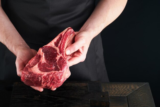 Lo chef taglia la bistecca di manzo Le mani dell'uomo tengono la bistecca cruda Tomahawk su un tagliere di legno rustico su sfondo nero Ricette di cucina e concetto di alimentazione Focus selettivo