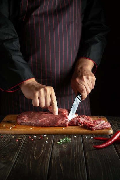 Lo chef taglia la carne di manzo cruda su un tagliere da cucina prima di grigliare Idea per un menu di hotel su sfondo scuro