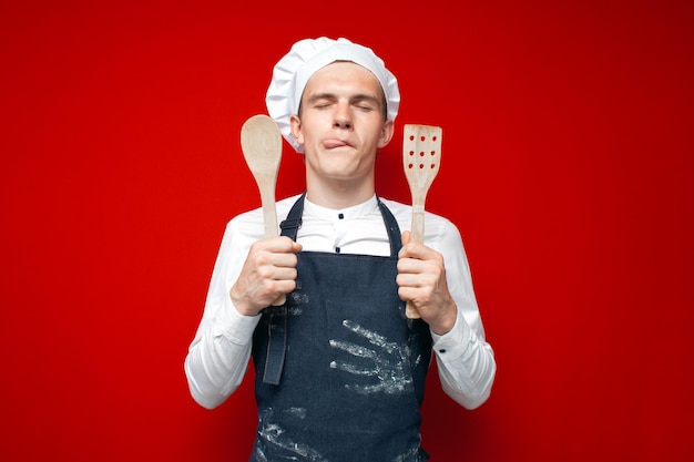 Lo chef tiene i cucchiai e si lecca le labbra su sfondo rosso isolato divertente lavoratore di cucina in uniforme