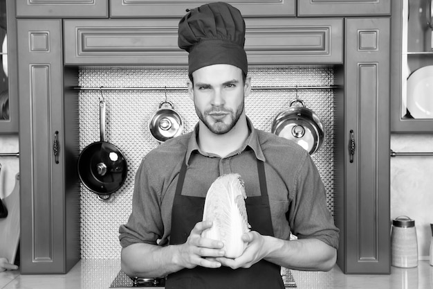 Lo chef uomo in grembiule con cappello rosso tiene verdura di cavolo cinese in cucina Concetto di dieta vegetariana per la salute Preparazione del cibo cucina cucina culinaria