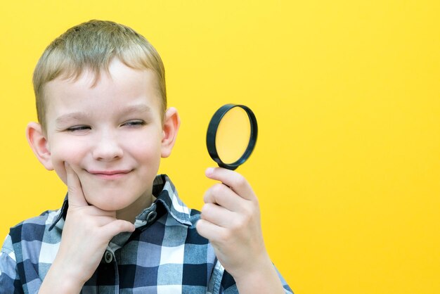 Lo scolaro curioso positivo in abiti casual guarda la telecamera attraverso una lente d'ingrandimento su uno sfondo giallo sorpresa