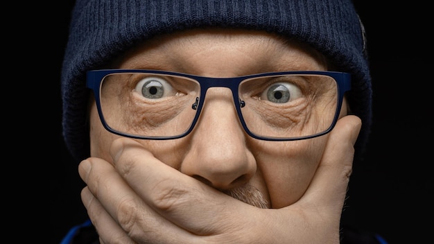 Lo sguardo sorpreso e spaventato di un uomo di mezza età con gli occhiali e un cappello lavorato a maglia blu Mano su fa