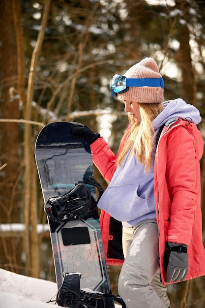 Lo snowboarder della donna caucasica tiene lo snowboard sul bellissimo sfondo della foresta innevata in una giornata di sole