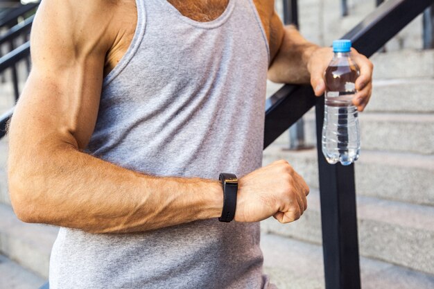 Lo sportivo con una bottiglia d'acqua sta riposando e controllando il suo orologio intelligente dopo aver corso