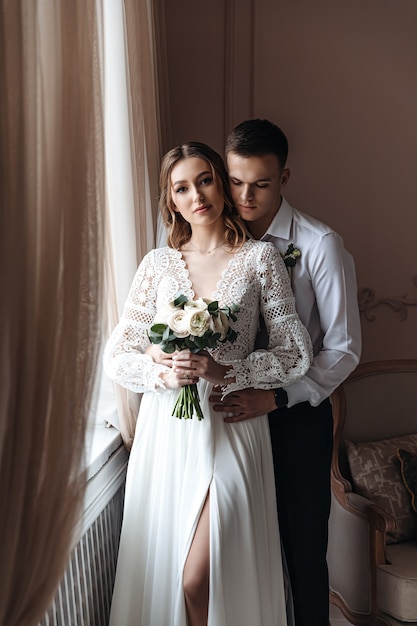 Lo sposo abbraccia dolcemente la sposa in un bellissimo abito di pizzo con un bouquet di fiori freschi. Sessione fotografica di matrimonio