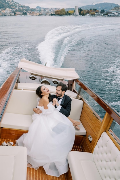 Lo sposo abbraccia la sposa che si appoggia all'indietro mentre è seduto in grembo su una barca a motore che naviga sul lago Como
