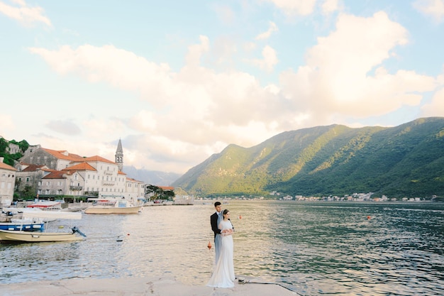 Lo sposo abbraccia la sposa da dietro mentre si trova sul molo sullo sfondo delle montagne della baia
