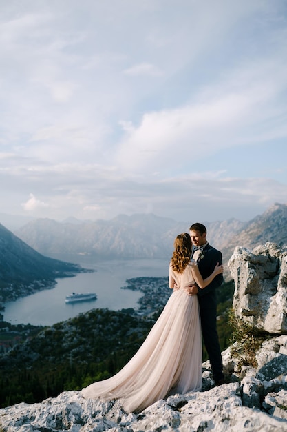 Lo sposo abbraccia la sposa per la vita su una montagna rocciosa che domina la baia