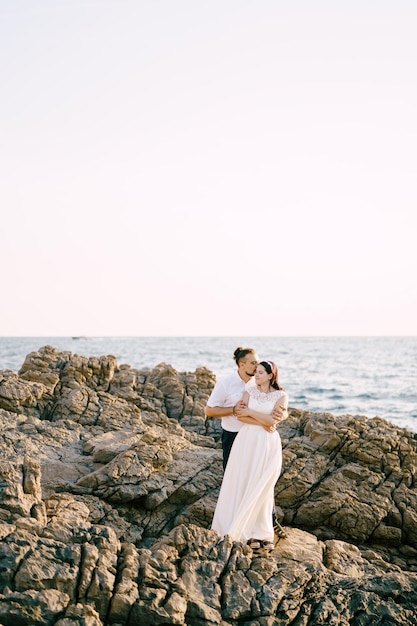 Lo sposo abbraccia la sposa per le spalle mentre si trova sulla costa rocciosa in riva al mare