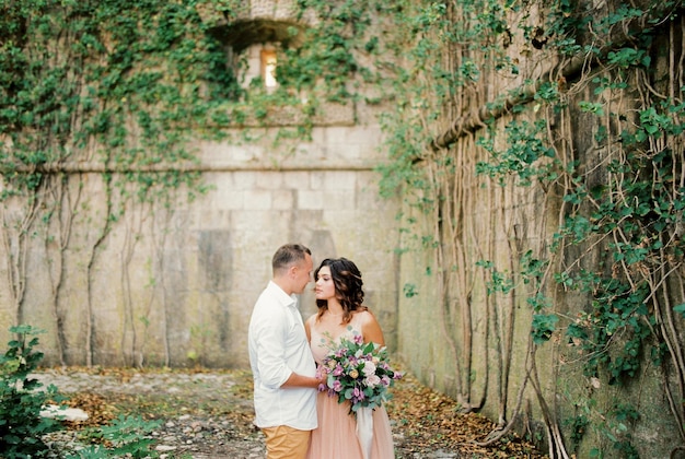 Lo sposo abbraccia la sposa vicino al muro di pietra ricoperto di edera verde