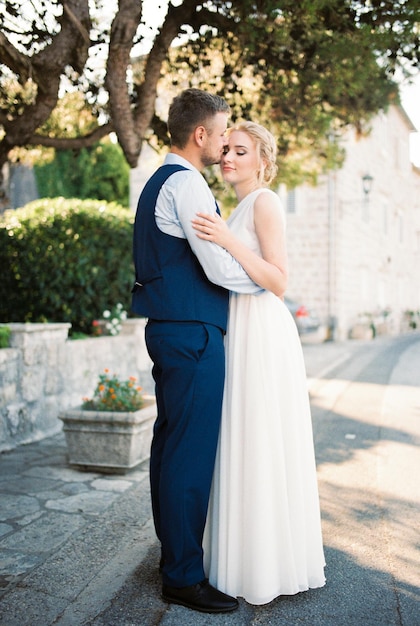 Lo sposo bacia la sposa sulla fronte abbracciandola alla vita vicino all'albero