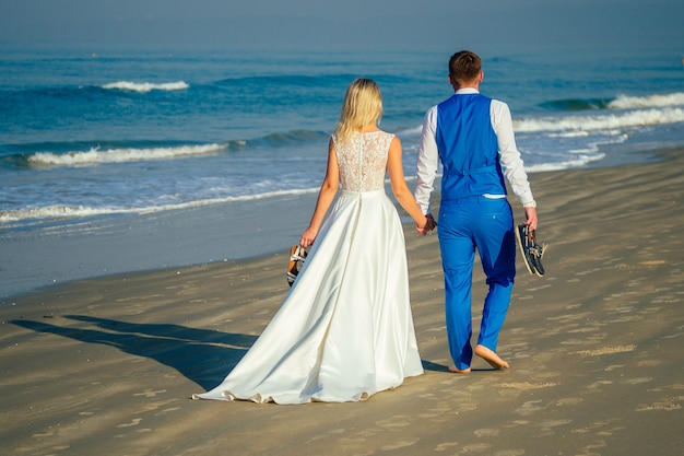 Lo sposo bello in un vestito elegante e una bella sposa in un abito da sposa camminano sulla spiaggia (vista dal retro). concetto di una cerimonia di matrimonio chic e ricca sulla spiaggia