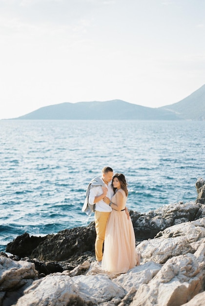 Lo sposo con una giacca sulla spalla abbraccia la sposa su una spiaggia rocciosa