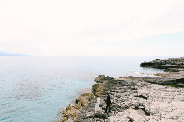 Lo sposo e la sposa camminano lungo la costa rocciosa vicino al mare tenendosi per mano