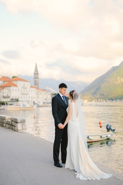 Lo sposo e la sposa si tengono per mano mentre sono in piedi sul molo sullo sfondo delle barche della baia e