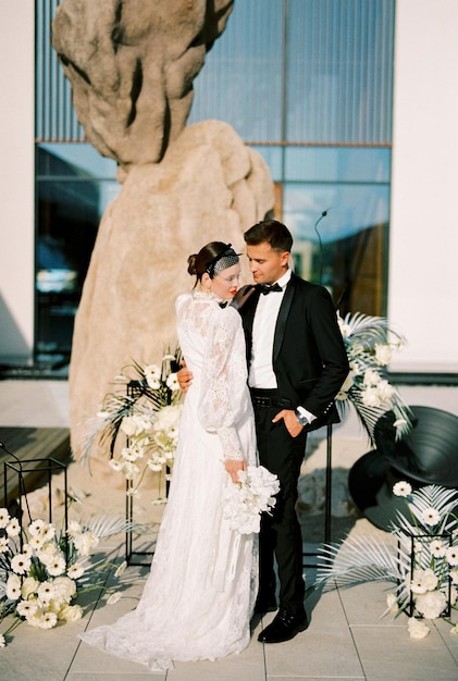 Lo sposo guarda la sposa che le abbraccia la vita mentre si trova vicino al semiarco nuziale di bouquets di