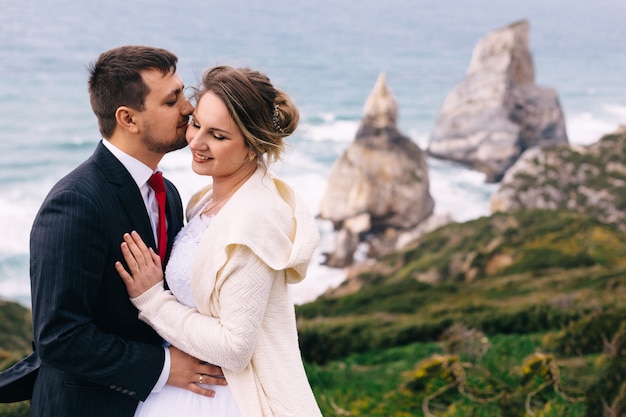 Lo sposo in abito con cravatta bacia la sposa in abito da sposa e maglione. gli sposi si abbracciano e chiudono gli occhi. vista sull'oceano e sulle scogliere.