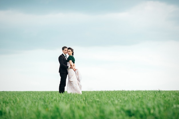 Lo sposo in abito marrone e la sposa in abito color avorio su un campo verde che si allontana in lontananza contro il cielo