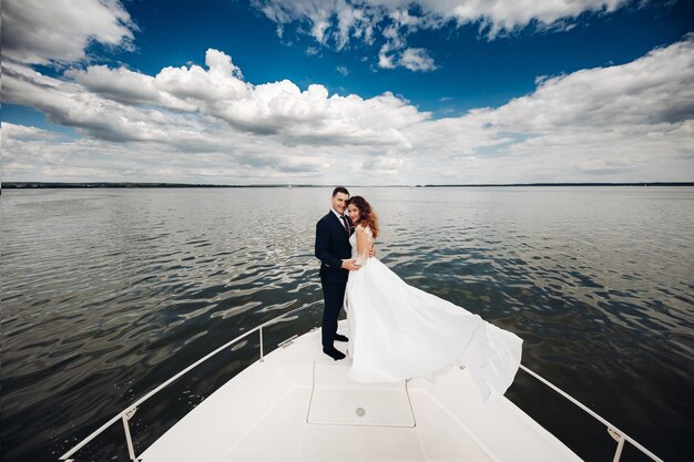 Lo sposo in abito nero con la sposa in abito lungo bianco cavalca insieme su un grande yacht bianco