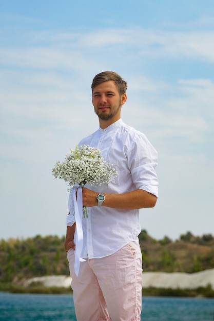 lo sposo in camicia bianca con un mazzo di fiori bianchi si trova vicino al lago blu