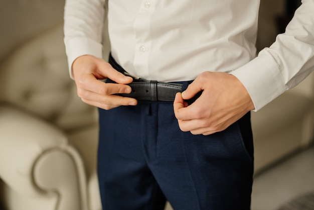 Lo sposo in pantaloni blu e una camicia bianca gli allaccia la cintura di pelle. Sposo del mattino. Dettagli del matrimonio. Concetto e composizione. Uomo d'affari e affari.