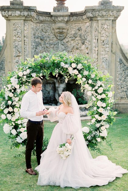 Lo sposo mette un anello al dito della sposa in abito bianco all'arco nuziale in giardino