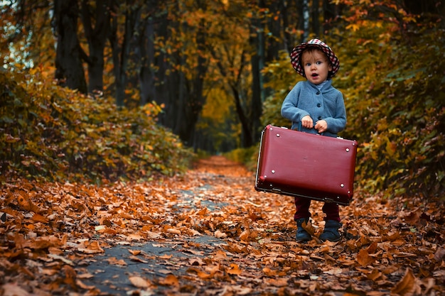 Lo stile retrò del bambino tiene una vecchia valigia sullo sfondo di un parco autunnale e foglie secche